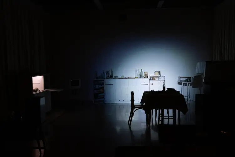 A darkened gallery room: a tiled kitchen counter lit pale blue, crowded with bottles and jars, a table and chairs in shadow before it.