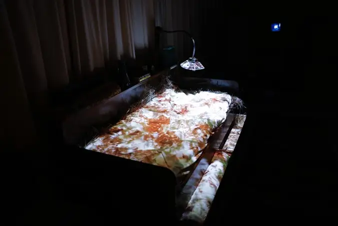 A bed with a projection of warm orange and white forms cast across the bedding, a lamp bent low over it.